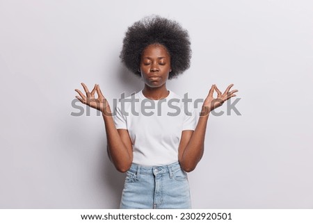 Horizontal shot of dark skinned woman keeps calm in yoga pose makes zen nirvana gesture meditates with closed eyes wears casual t shirt and jeans isolated over white background. Stay always relaxed