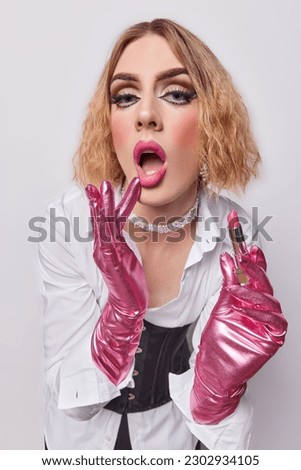 Indoor shot of gay with vivid makeup keeps mouth widely opened holds lipstick wears elegant outfit gets prepared for date with boyfriend isolated on white background. Transgender woman applies makeup