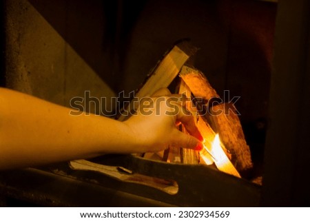 Female hand holding a burning match reaches for the wood in a fireplace to light up a fire and keep a log country house warm from inside during a cold season. Autumn or winter evening at cozy home.  Royalty-Free Stock Photo #2302934569