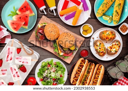 Summer BBQ food table scene. Hamburgers, hot dogs, potatoes, corn and cold treats. Above view over a dark wood background.