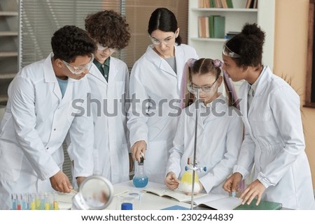 Diverse group of young teens wearing lab coats doing science experiments in school with teacher helping Royalty-Free Stock Photo #2302944839