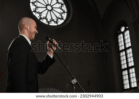 Mature pastor in black suit speaking prayer in microphone while standing in old baptist church Royalty-Free Stock Photo #2302944903