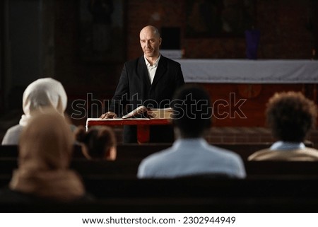 Mature preacher standing at altar with Bible and reading prayer for people in church Royalty-Free Stock Photo #2302944949