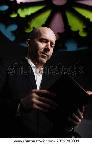 Vertical image of mature man in black suit reading Bible standing in old baptist church Royalty-Free Stock Photo #2302945001