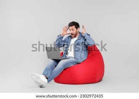 Emotional man with laptop sitting on beanbag chair against light grey background