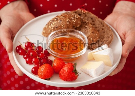 The girl in a red dress holds a plate with whole grain bread, cheese, apricot jam, red currants and strawberries. Healthy lifestyle