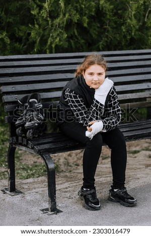A schoolgirl girl in sadness, a child athlete with a plaster on her arm after a bone fracture sits on a bench in the park with roller skates, missing the ride. Photography, portrait, sport concept.