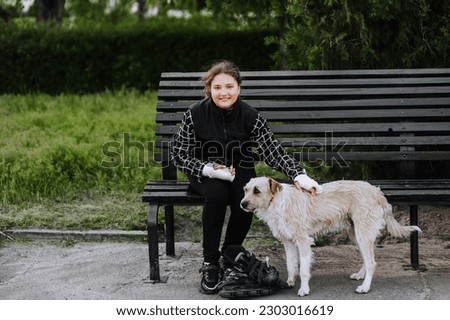 A girl in sadness, a child athlete with a cast, a bandage on her hands after a bone fracture, sits on a bench in the park with roller skates and plays a mongrel dog. Photography, portrait, sports.