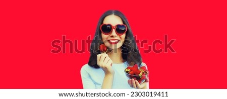 Portrait of happy smiling woman with handful of fresh strawberries wearing red heart shaped sunglasses on background