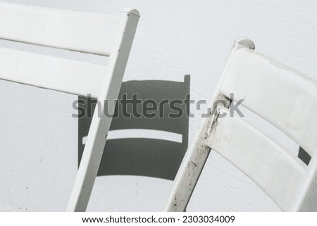 A detail shot of two folding white lawn chairs on a hot, bright day, with harsh sun shadows, against a white background