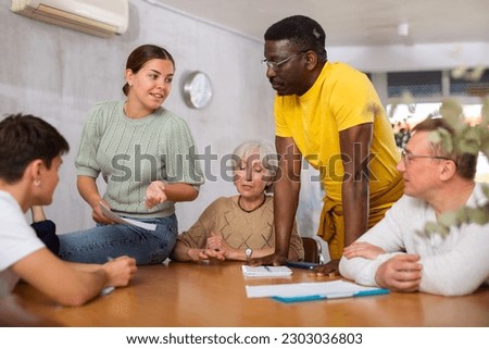 Interested friendly young girl, student of adult school, taking part in discussion of study topics with international mixed age group of coursemates sitting at table.. Royalty-Free Stock Photo #2303036803