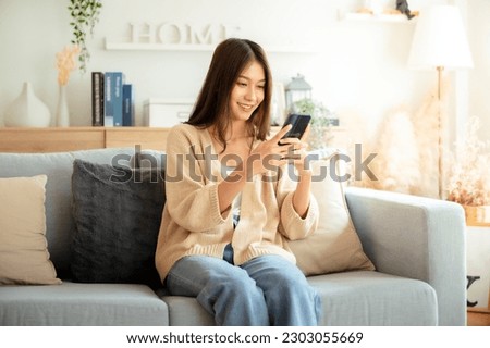 Happy young asian woman relax on comfortable couch at home texting messaging on smartphone, smiling girl use cellphone chatting, browse wireless internet on gadget, shopping online from home Royalty-Free Stock Photo #2303055669