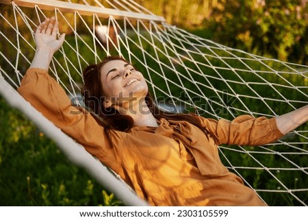 a happy woman is resting in a hammock with her eyes closed and her hands behind her head smiling happily enjoying the day
