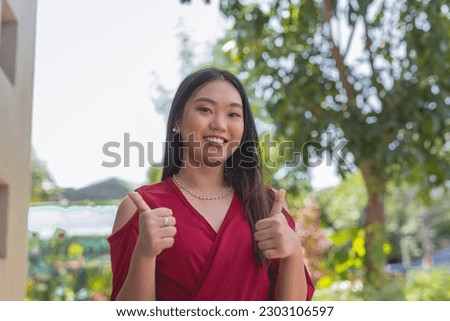 A close- up picture of a lovely Asian woman, looking sweetly at the camera, smiling widely and holding up a two thumbs up sign. A tree is shown in the background.