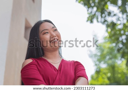 A close- up picture of a lovely white Asian woman, arms crossed while gazing far away and smiling widely. A tree and a building is shown in the background.