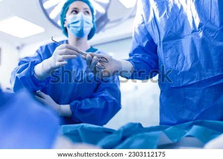 Low Angle Shot in the Operating Room, Assistant Hands out Instruments to Surgeons During Operation. Surgeons Perform Operation. Professional Medical Doctors Performing Surgery. Royalty-Free Stock Photo #2303112175