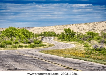 A tranquil landscape of a cloud-filled sky over an expansive dam, tree-lined roads and plants in San Angelo State Park, Texas. Royalty-Free Stock Photo #2303112801