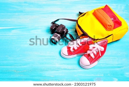 Set of small red high-top canvas shoes, a tiny backpack, and a toy camera arranged on a blue wooden background. Creative back-to-school concept with the essentials for a child heading back to class.