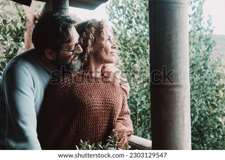 Adult couple in love and relationship looking outside home enjoying relax and serene moment in terrace house patio together hugging and loving. Healthy nature lifestyle people. Alternative wooden home Royalty-Free Stock Photo #2303129547