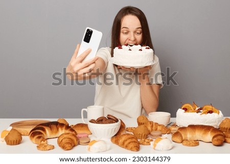 Funny woman blogger wearing white T-shirt isolated over gray background biting big cake biting dessert taking selfie creating content celebrating eating cheat meal.