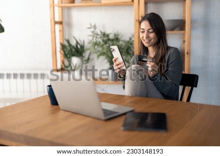 Happy young woman, satisfied online buyer holding credit card, using e-banking app on smart mobile phone making convenient financial e-commerce payment digital transaction while sitting at home. Royalty-Free Stock Photo #2303148193
