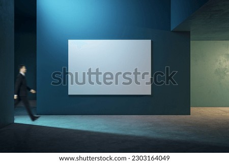 Blurred businessman walking by big white blank poster with place for your logo or text on dark wall background in abstract hall area with concrete floor, mockup