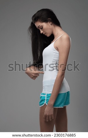 Beautiful young woman with long hair, white tank top, blue sporty shorts, worries about her belly, pointing at it. Some girls never like themselves despite their beauty. Doubts about diet and thinness