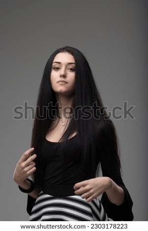 Long-haired girl, elegantly dressed, arms in motion. Haughty, snobbish look, very beautiful in black top and black-and-white skirt. She is fashion personified. Gray background. Very serious Royalty-Free Stock Photo #2303178223