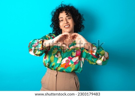 young arab woman wearing colorful shirt over blue background smiling in love doing heart symbol shape with hands. Romantic concept.