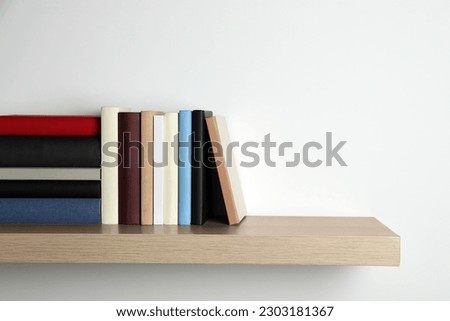Many hardcover books on wooden shelf near white wall. Space for text