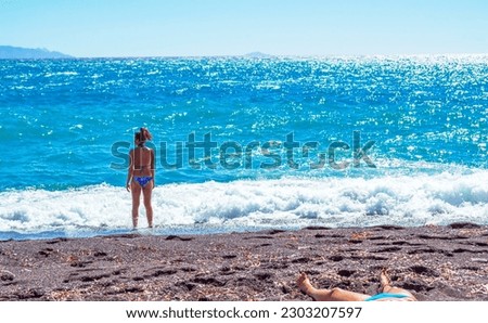 Sunbathing woman in a swimsuit on the seashore enjoying the amazing Aegean Sea vista.Picture taken in Kamari,Santorini,Greece- unique,amazing island.In the distance seen Anafi island.close to nature,