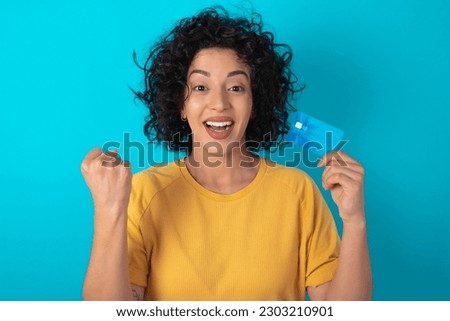 Photo of lucky impressed young arab woman wearing yellow T-shirt over blue background arm fist holding credit card. Celebrated