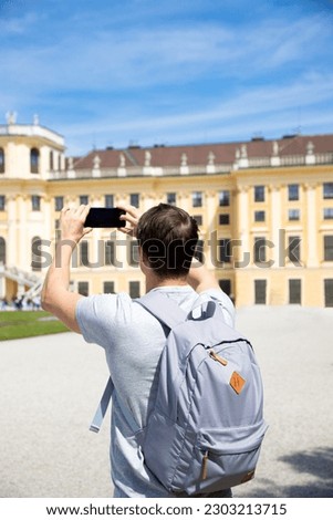 A young man takes pictures of the sights of Austria on his smartphone. A warm and sunny summer day. back view. The guy is dressed in ordinary clothes and a gray backpack.