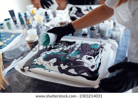 Master class on an Acrylic Fluid Pouring. Teacher and teen girl paint with liquid acrylic in an art workshop. Fluid Art. Dirty glass technique. There are paintings, paints and brushes on the table Art Royalty-Free Stock Photo #2303214031