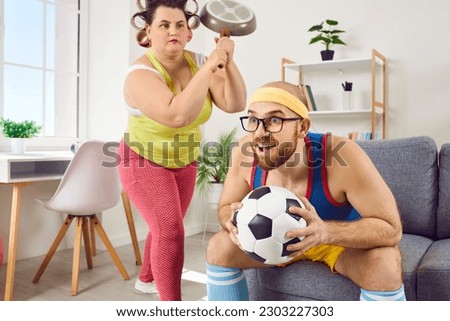 Funny angry frustrated evil housewife overwhelmed by bad mood emotions raises culinary skillet pan over head of happy lazy silly husband watching sport on TV, having fun and not helping with housework Royalty-Free Stock Photo #2303227303
