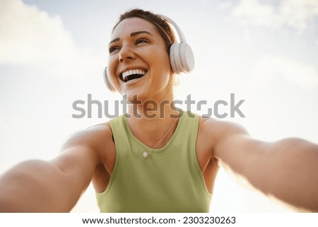 Fitness woman taking a fun selfie against the sky. Happy sports woman wearing wireless headphones. Woman having fun during her morning workout outdoors.