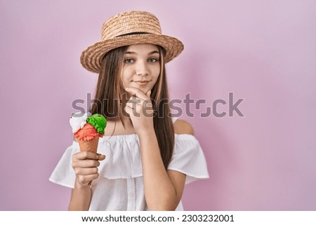 Teenager girl holding ice cream looking confident at the camera smiling with crossed arms and hand raised on chin. thinking positive. 
