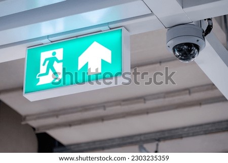 Fire exit sign with closed-circuit television (CCTV) camera installed on ceiling structure in the building. Security and safety system for public building