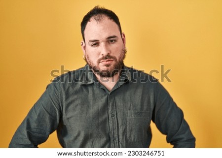 Plus size hispanic man with beard standing over yellow background looking sleepy and tired, exhausted for fatigue and hangover, lazy eyes in the morning. 