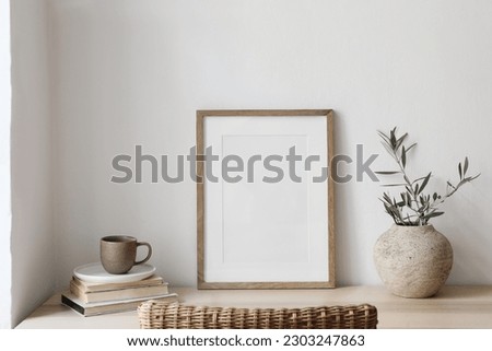 Closeup of picture frame, poster mockup. Vase with olive tree branches on wooden table. Blurred rattan chair. Cup of coffee, books. Summer artistic Mediterranean interior. Working space, home office. 