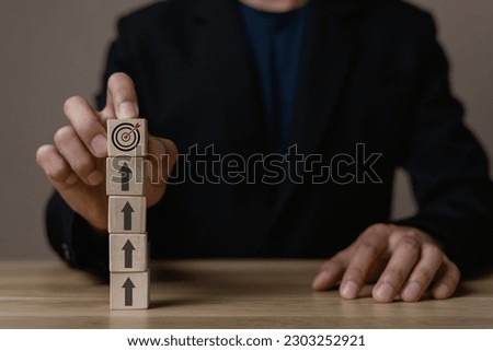 Businessman holding wooden cube with target board icon and arrow on wooden table. Goals and planning for success in marketing business, achieve the objective concept. Closeup and free copy space.