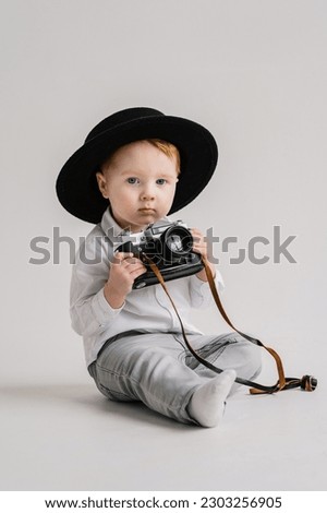 Child in a hat sitting on floor and play with photo camera. Fun kid holds retro vintage camera isolated on white wall. International Photographers Day. Children's studio portrait. Mockup. Close Up. Royalty-Free Stock Photo #2303256905