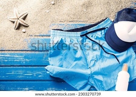 Flat lay shot of blue swim shorts with hat and suncream. Sand and starfish on blue weathered wooden beach platform.