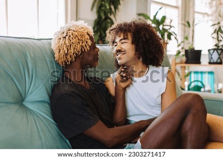 Young gay couple being romantic indoors. Two affectionate male lovers embracing and smiling at each other while sitting together in their living room. Young gay couple relaxing at home. Royalty-Free Stock Photo #2303271417
