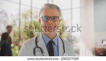 Portrait of a Middle Aged Medical Health Care Professional Posing, Smiling and Looking at Camera. Caucasian Clinic Physician Wearing Glasses and a White Coat Working in Hospital Office Royalty-Free Stock Photo #2303277339