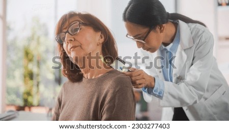 Portrait of a Young Asian Dermatologist Using a Medical Magnifying Glass to Inspect any Damages on the Skin of a Senior Patient During a Health Check Visit to a Clinic. Doctor Working in Hospital Royalty-Free Stock Photo #2303277403