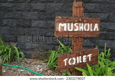 Wooden direction sign post to the mushola (prayer room) and toilet which is placed outdoor around green leaves and black stone wall. Wood material.