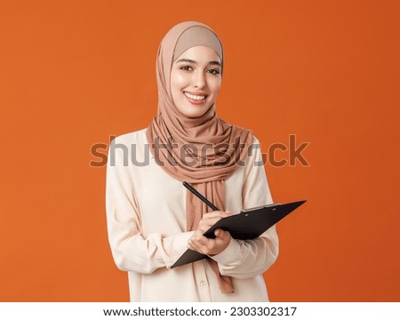 Beautiful woman in traditional muslim hijab writing notes on orange background
