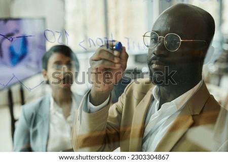 Focus on young serious African American businessman or coach with blue highlighter writing on transparent board during presentation Royalty-Free Stock Photo #2303304867