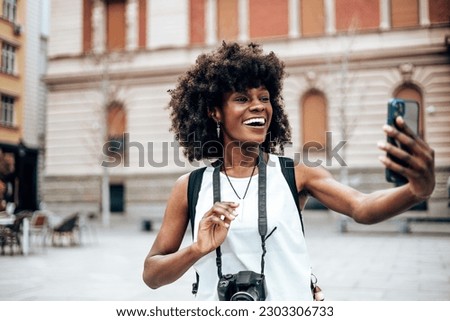 Young black female tourist enjoys walking through the streets of a beautiful European city. She is happy and using her smart phone to take some photos and videos. Bright sunny day. Royalty-Free Stock Photo #2303306733